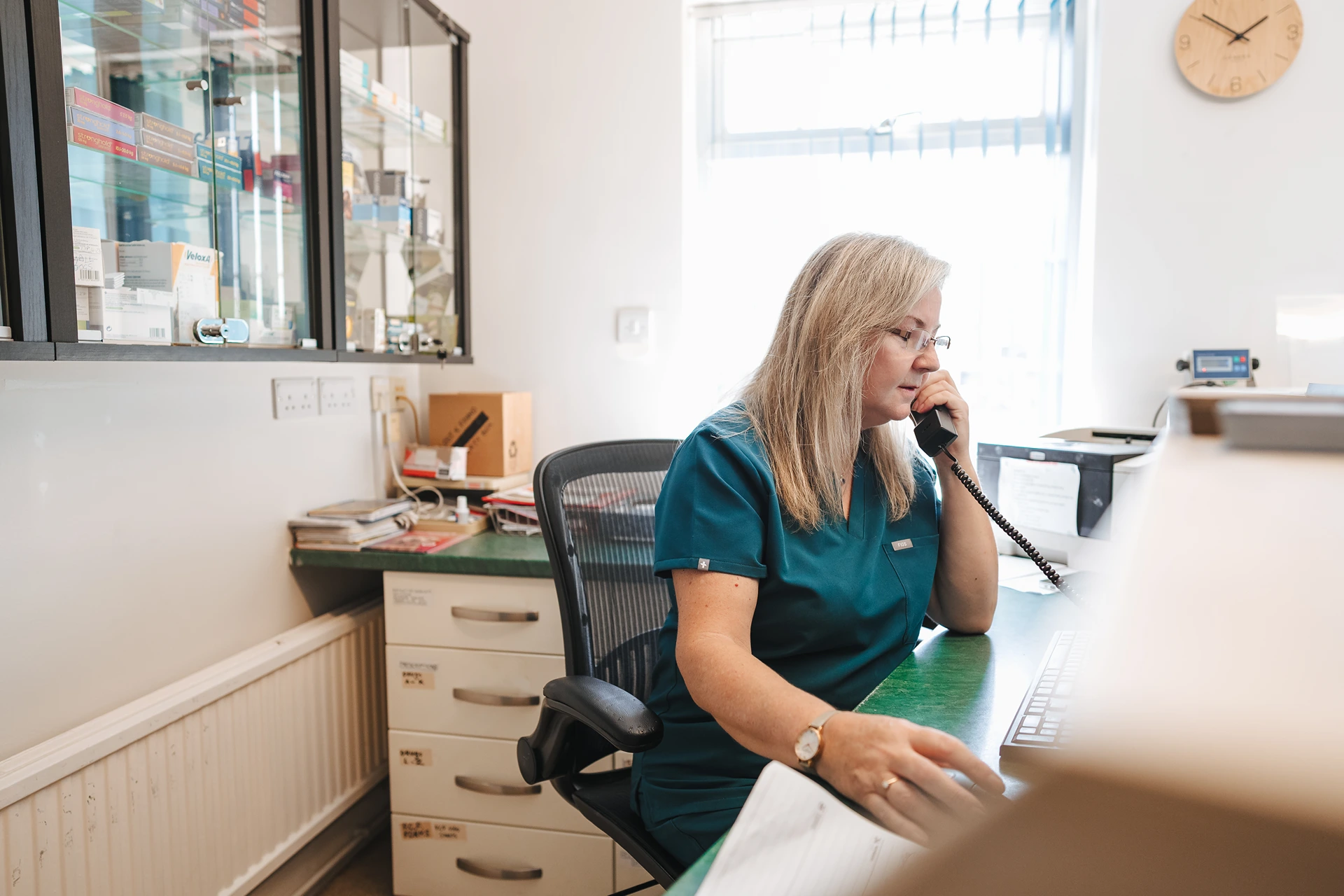 Woman in teal uniform sitting at a desk, talking on a phone in a bright office with shelves and a clock.