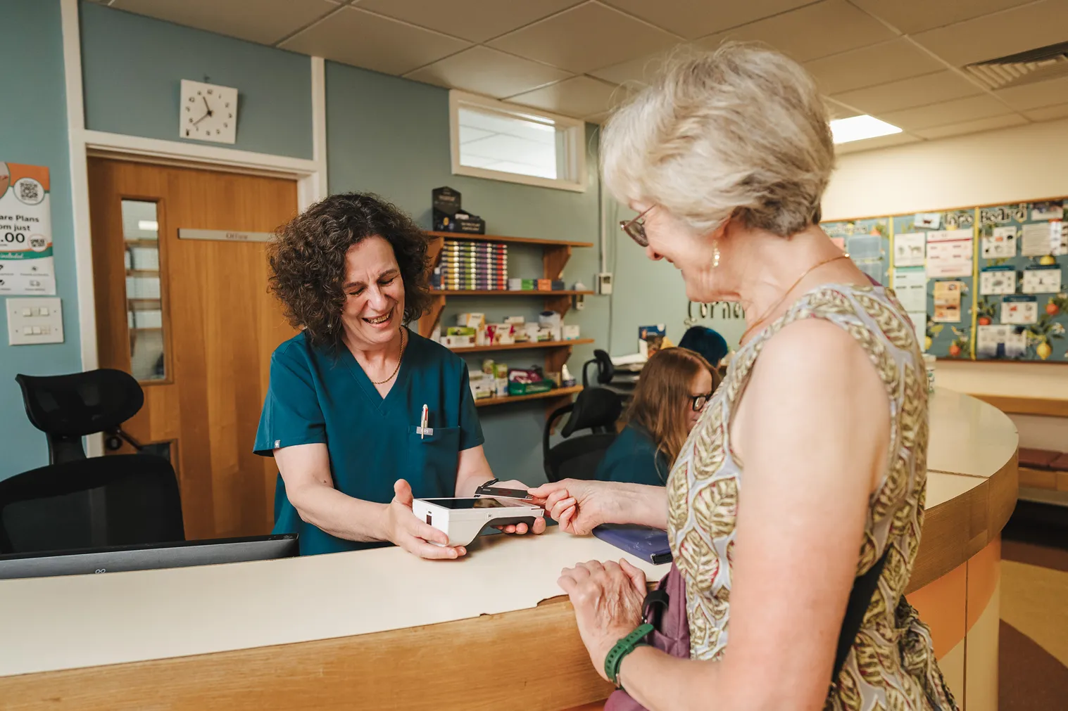 Healthcare worker in blue scrubs smiling as she holds a card reader for an elderly woman paying with a card.
