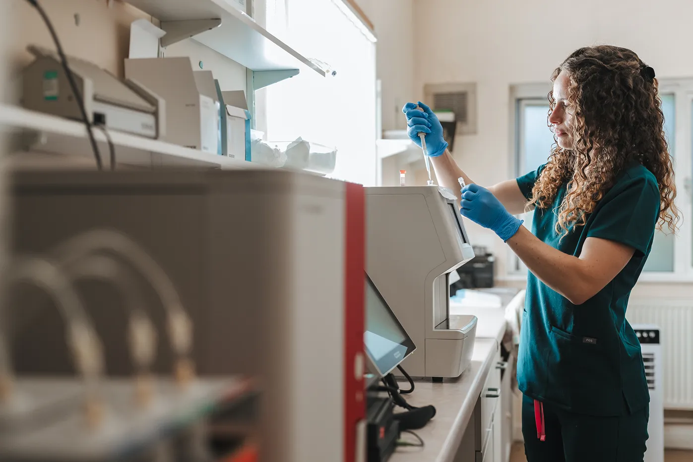 Woman in scrubs and gloves using a pipette with lab equipment in a bright laboratory.