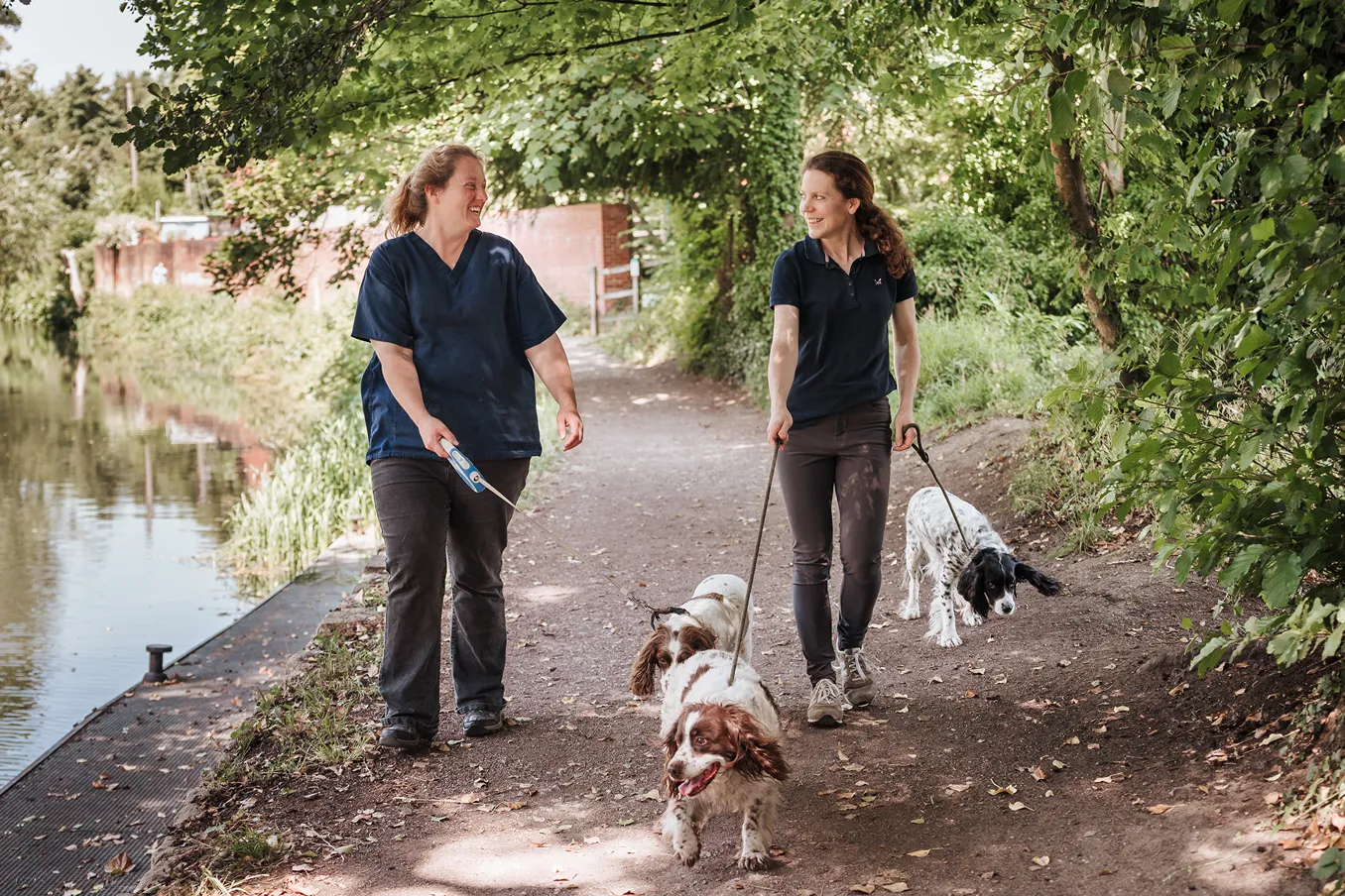 Two women walking three dogs on a dirt path beside a river under green trees.