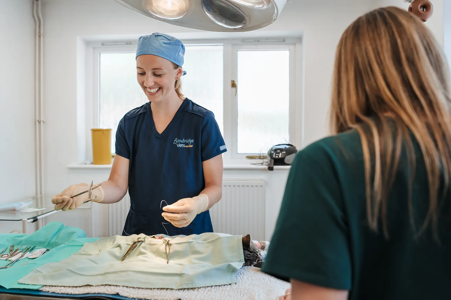 Veterinarian in blue scrubs smiling while preparing to suture in a clinical room with a covered animal