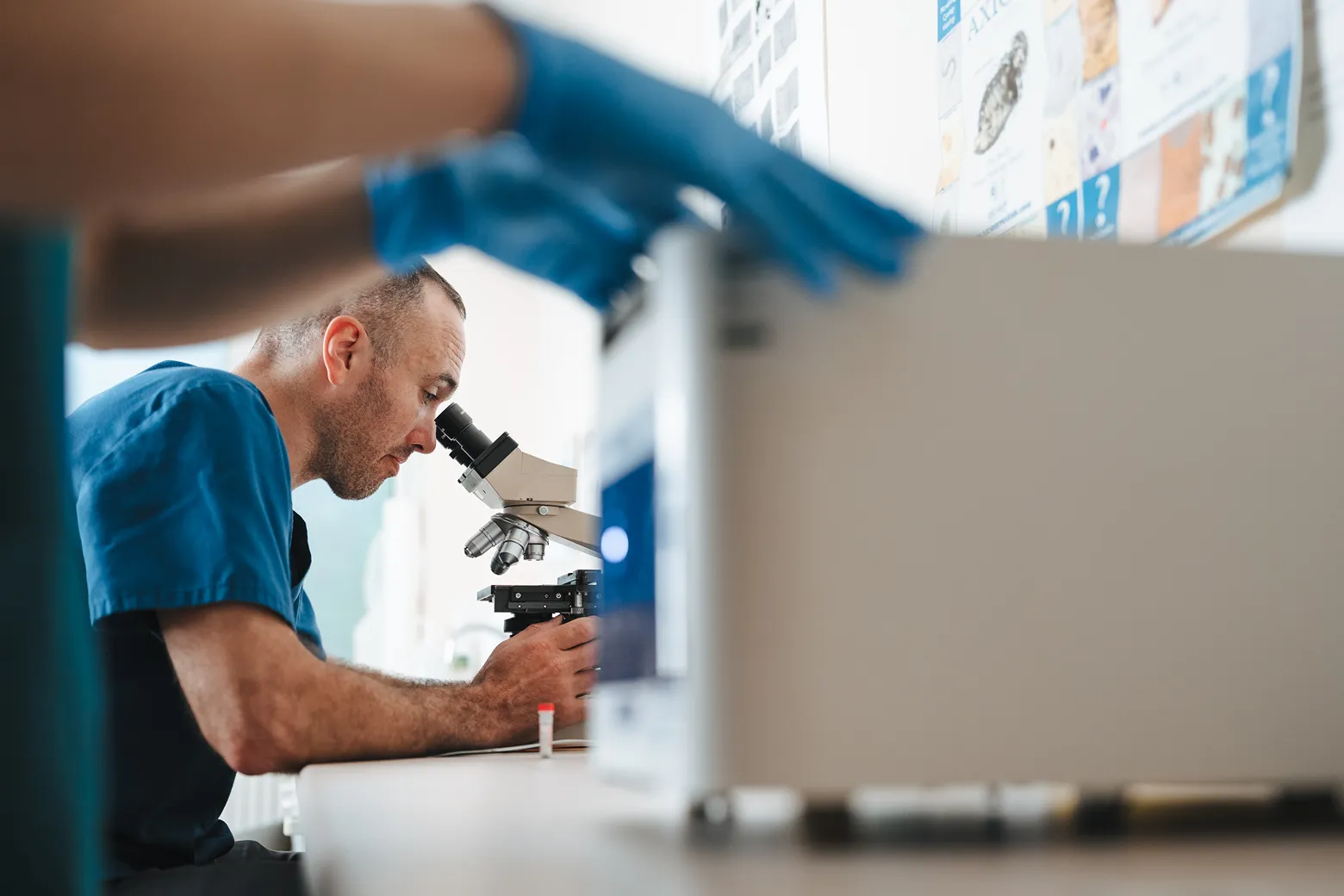 Scientist in blue scrubs examining a sample through a microscope in a lab setting.