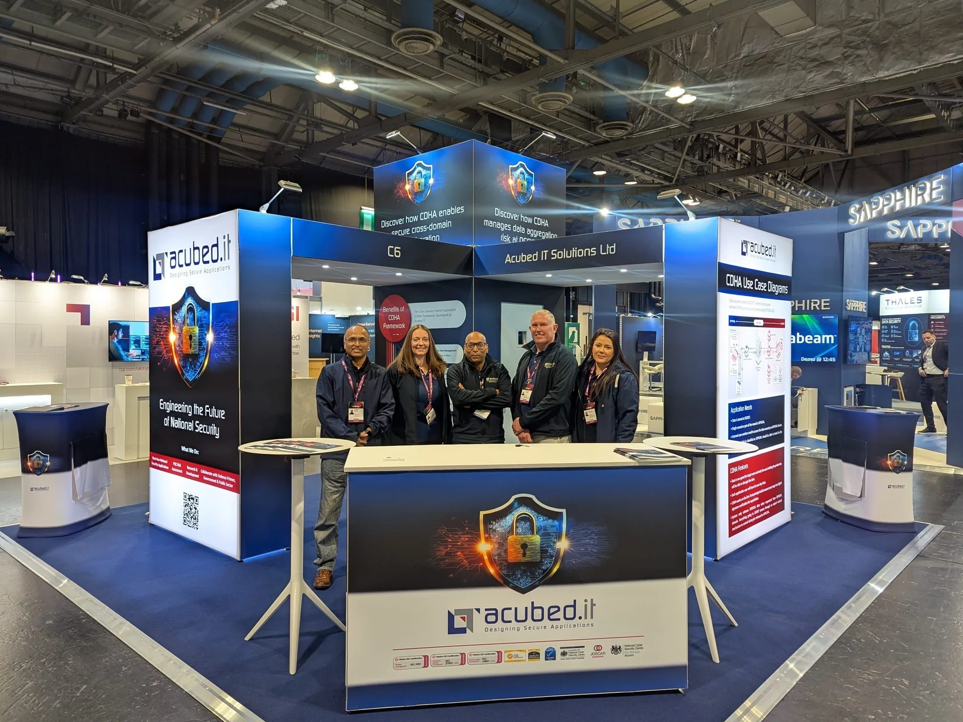 Acubed.IT stand at CYBERUK 2026: team behind the branded counter on a navy-carpeted stand with a graphic tower