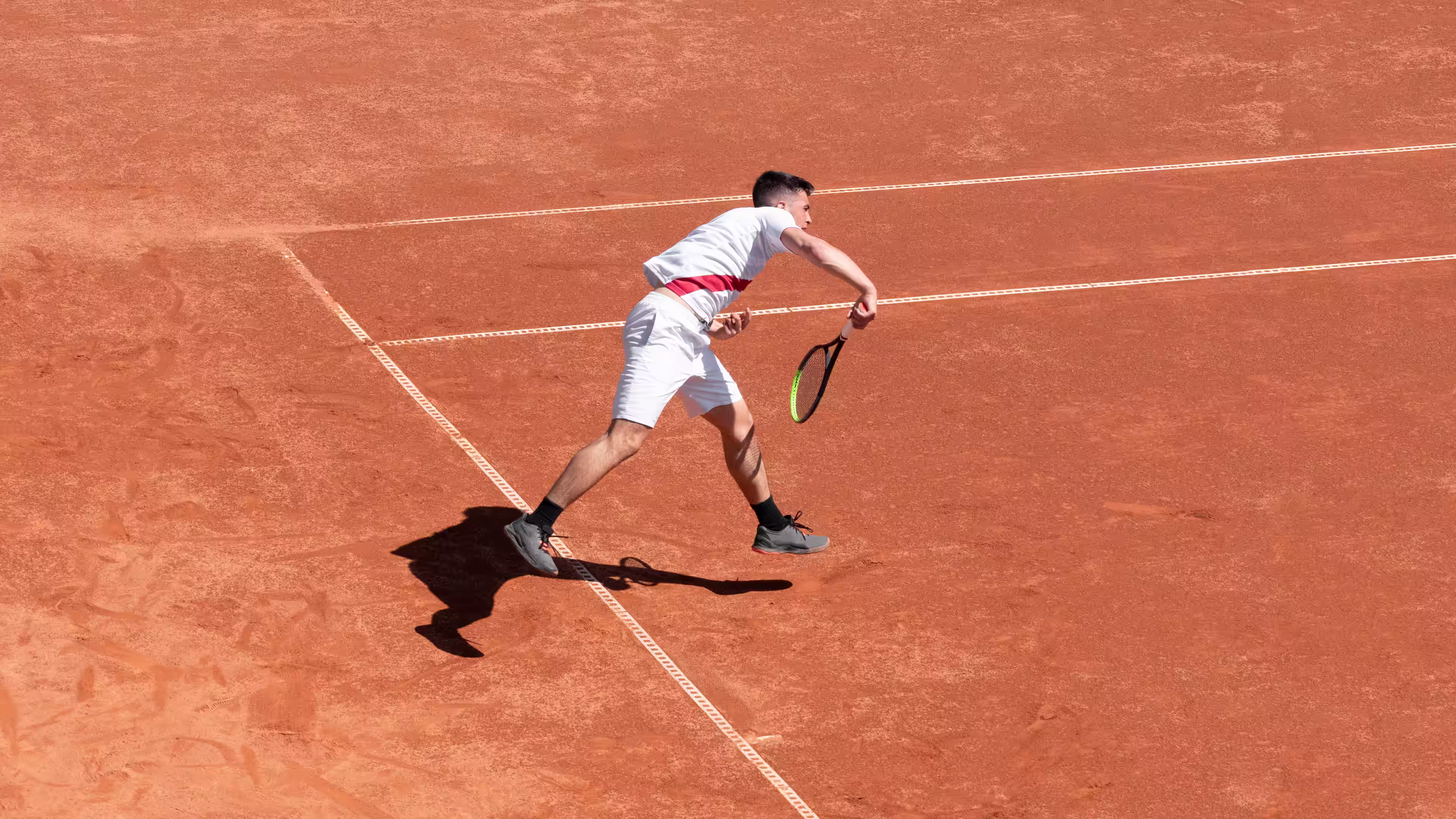 Tennis player in white completes serve with force lifting his feet off the clay court