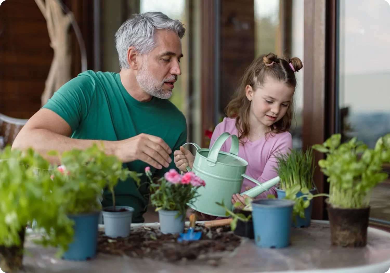 A man with gray hair and a beard, wearing a green shirt, helps a young girl in a pink shirt water potted plants on a table.