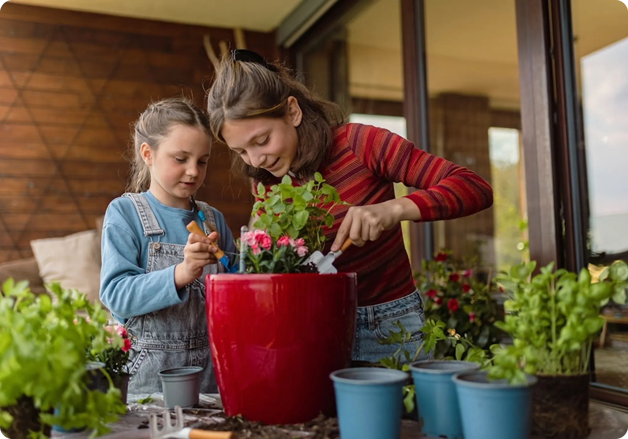 Two girls gardening together, using small tools to plant flowers in a red pot on a table with other plants nearby.