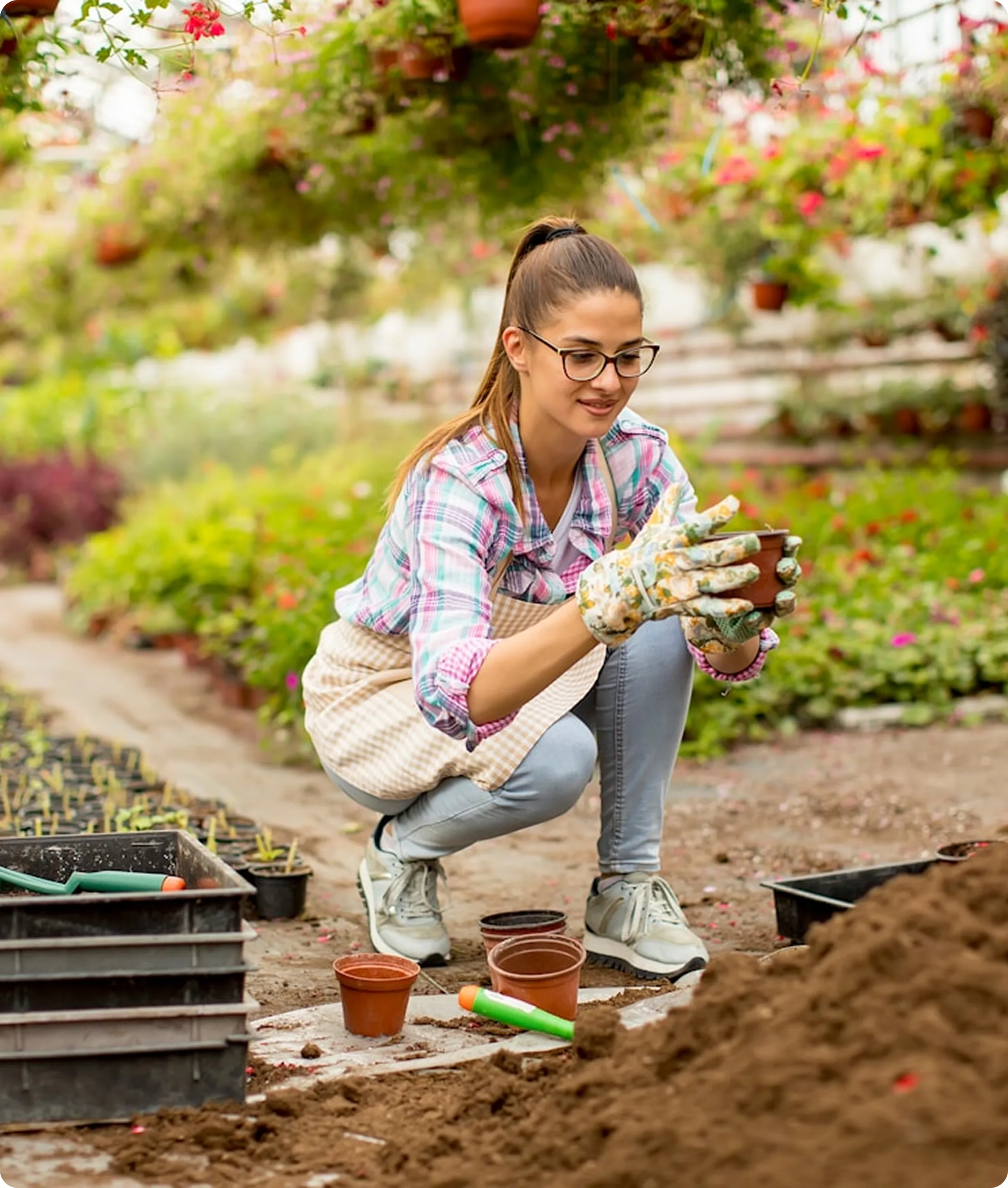 Woman wearing glasses and gloves crouching in a garden center, holding a small flower pot surrounded by gardening tools and pots.