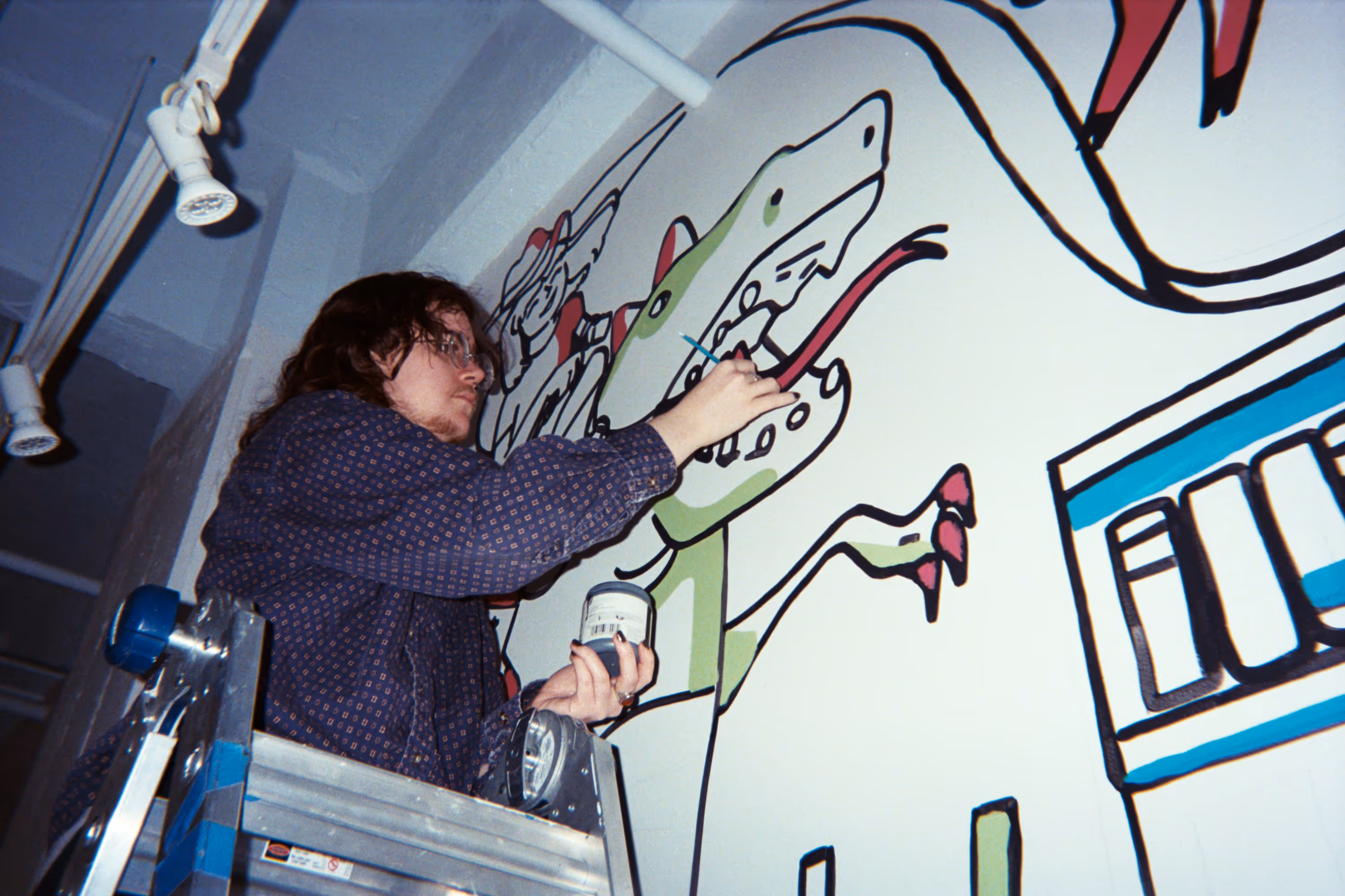 A medium close shot of a person with curly hair and glasses standing on a ladder painting a mural of a dinosaur on a white wall.
