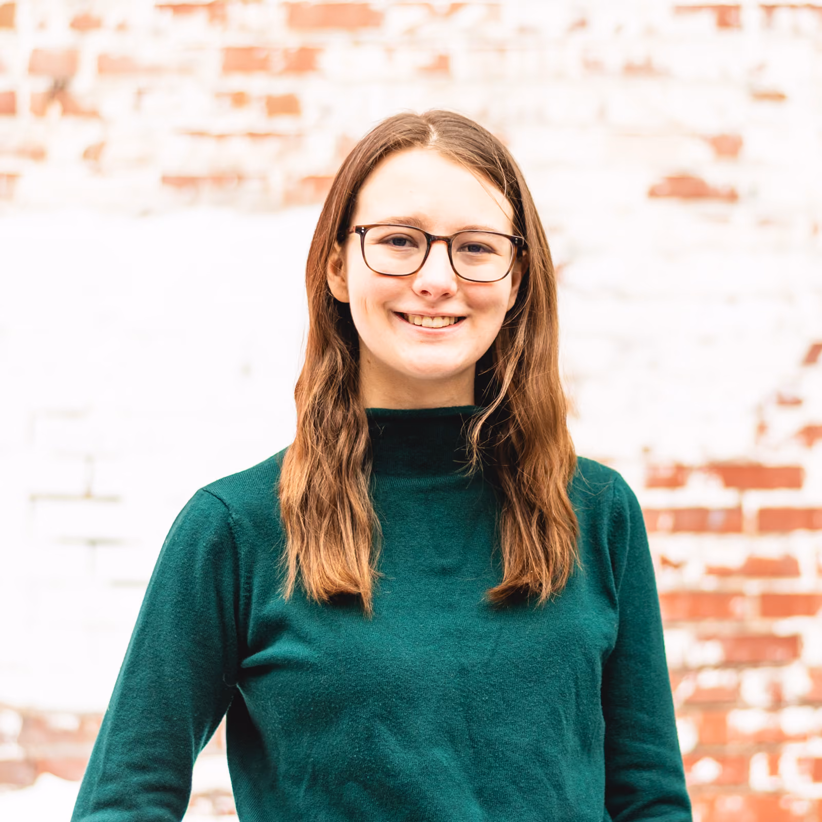 Portrait-style shot of a smiling young woman with medium wavy light-brown hair wearing a green turtle-neck sweater against a brick wall.