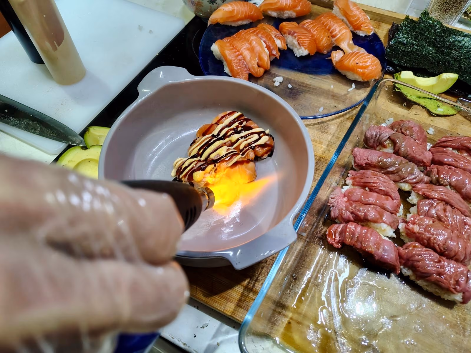 Flambee d'un sushi avec un chalumeau dans un bol gris, des sushis au saumon sur une assiette bleue et des sushis au bœuf sur une plaque en verre sur une surface en bois.