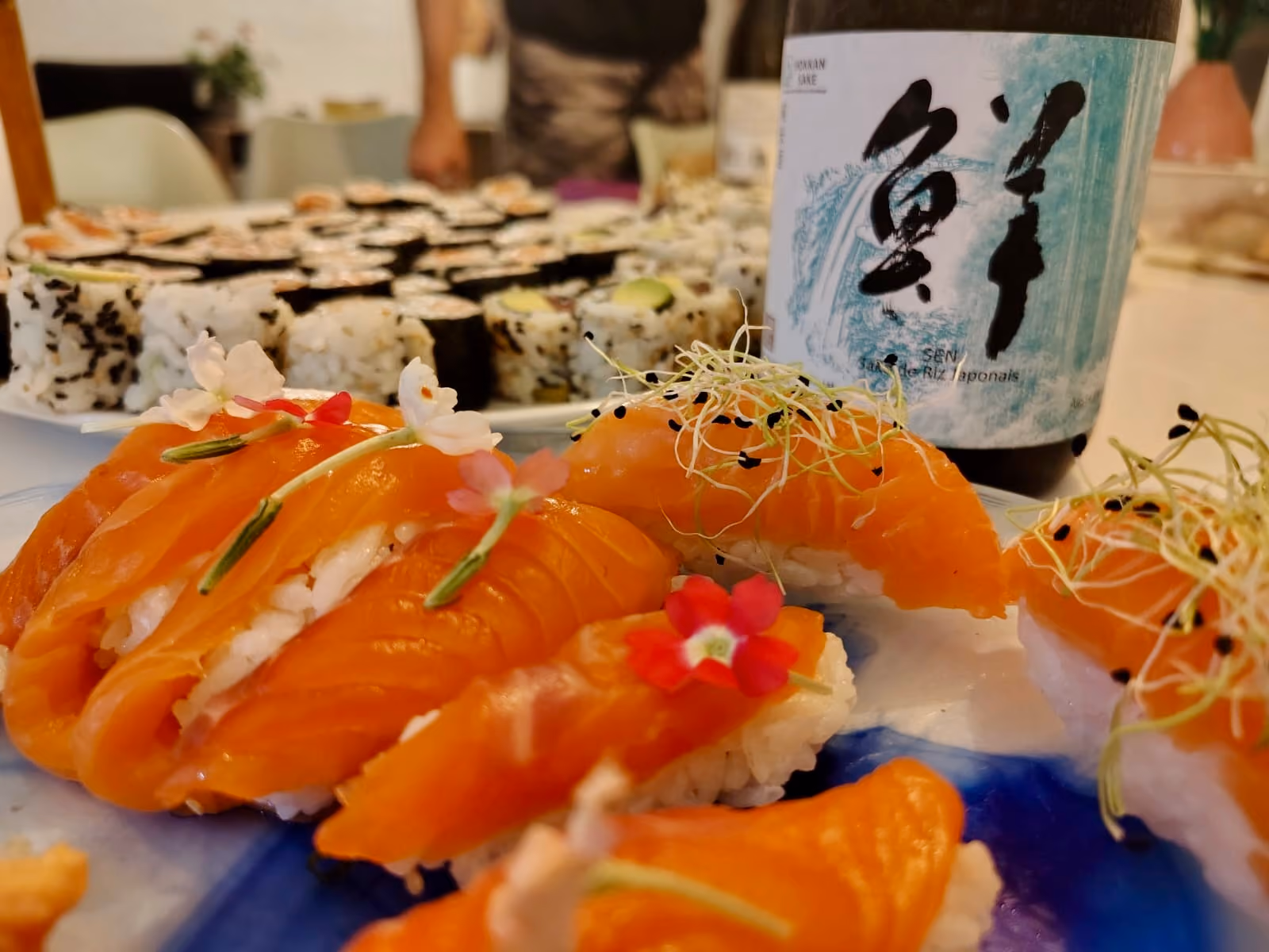 Assortiment de sushi avec des tranches de saumon décorées de petites fleurs et pousses, accompagné d'une bouteille de sake japonais en arrière-plan.