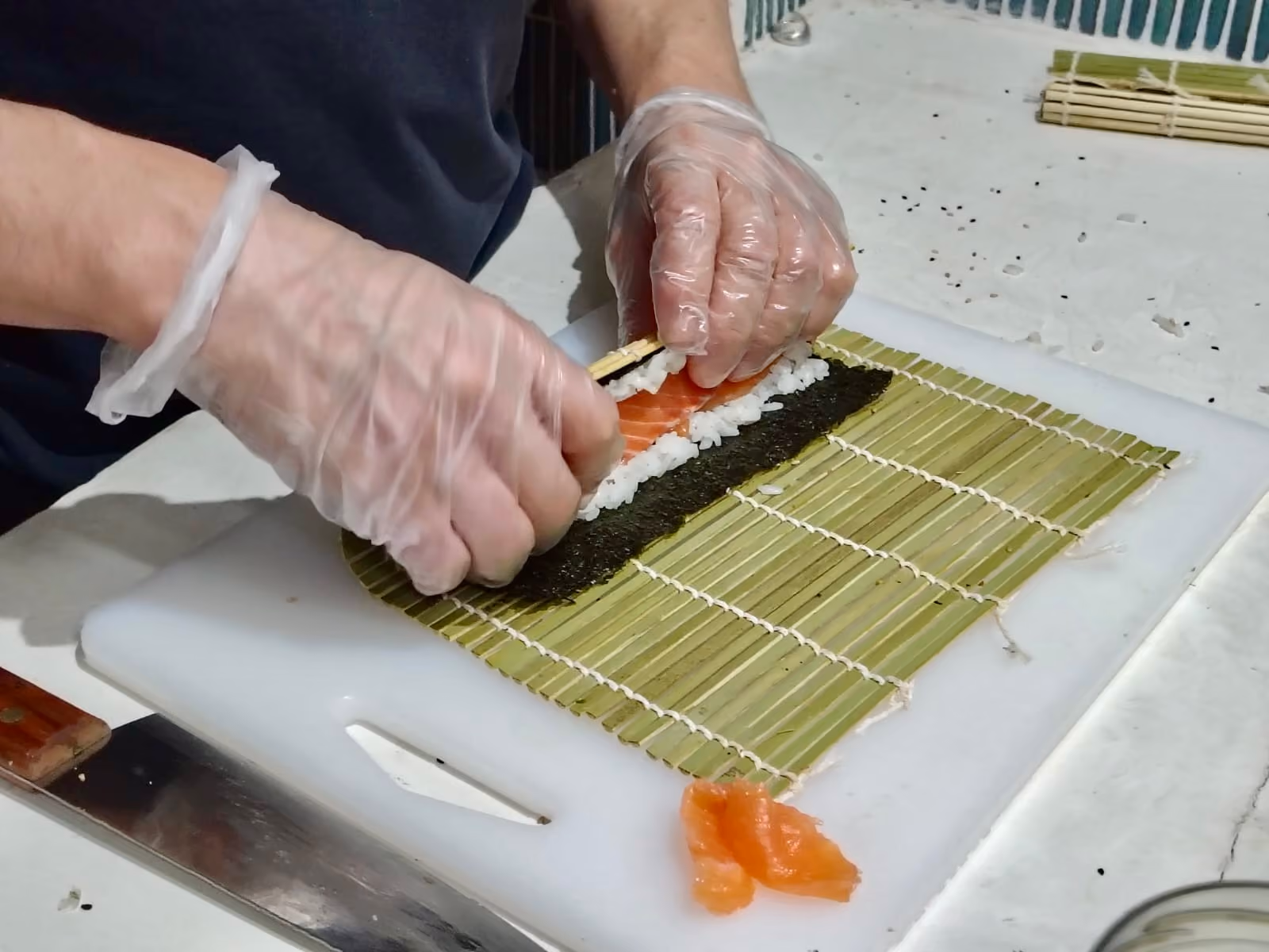 Une personne portant des gants en plastique prépare un maki en enroulant du saumon et du riz sur une feuille de nori posée sur un tapis en bambou.