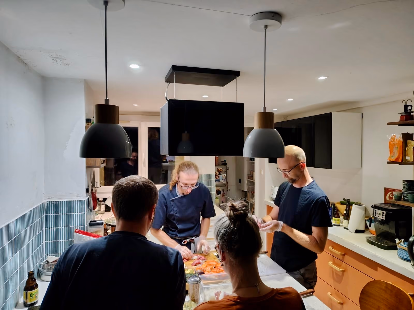 Quatre personnes préparant des sushis dans une cuisine moderne avec des suspensions au plafond, des meubles blancs et bleus, et des ustensiles de cuisine sur le comptoir.