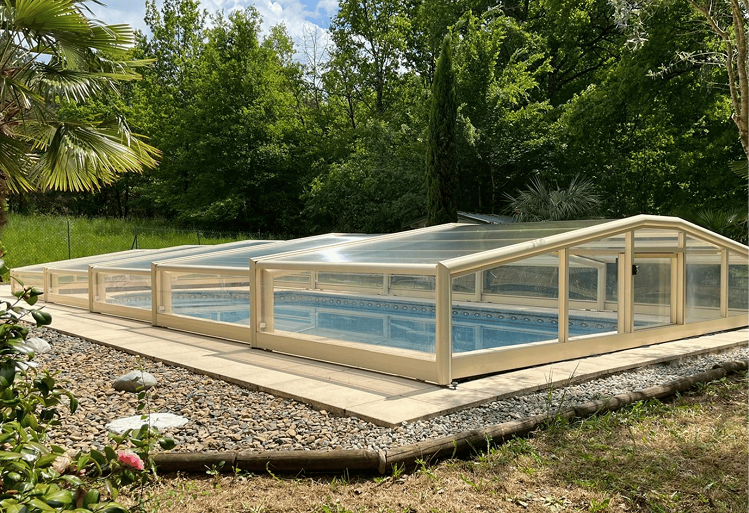 Piscine extérieure avec un abri transparent beige déployé, entourée de pierres décoratives et de végétation verte.