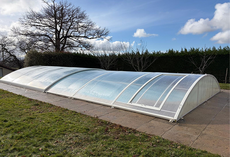 Une grande piscine couverte par une structure basse et translucide en plastique blanc, entourée d'une pelouse verte et entourée d'arbres et de buissons, sous un ciel bleu avec quelques nuages blancs.