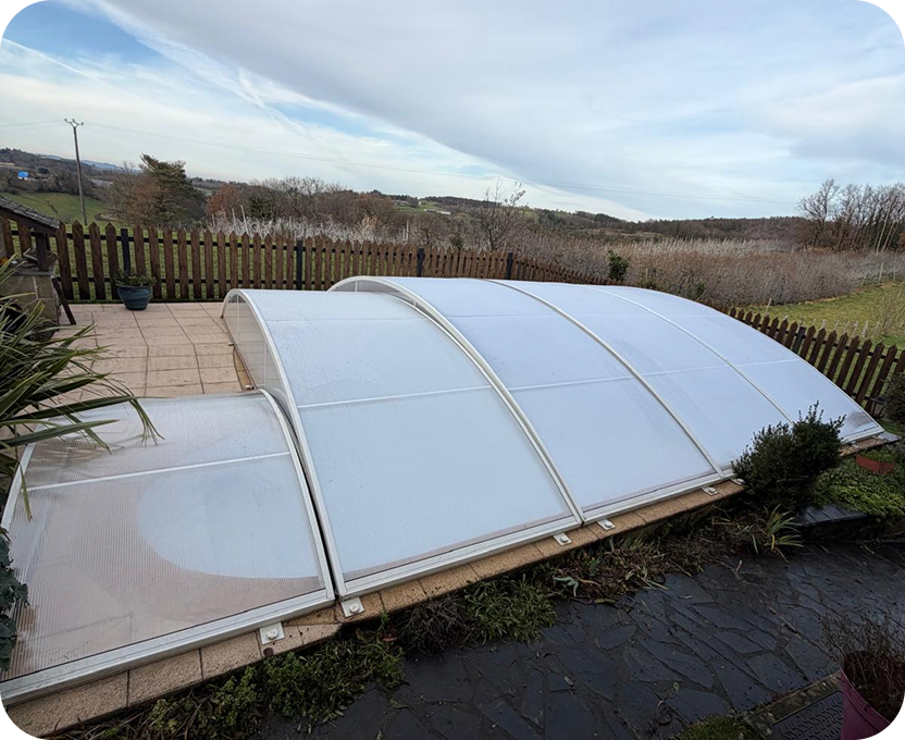 Piscine extérieure recouverte d'une structure de couverture transparente à plusieurs volets arrondis, entourée d'une terrasse pavée et d'une clôture en bois, avec un paysage rural en arrière-plan.