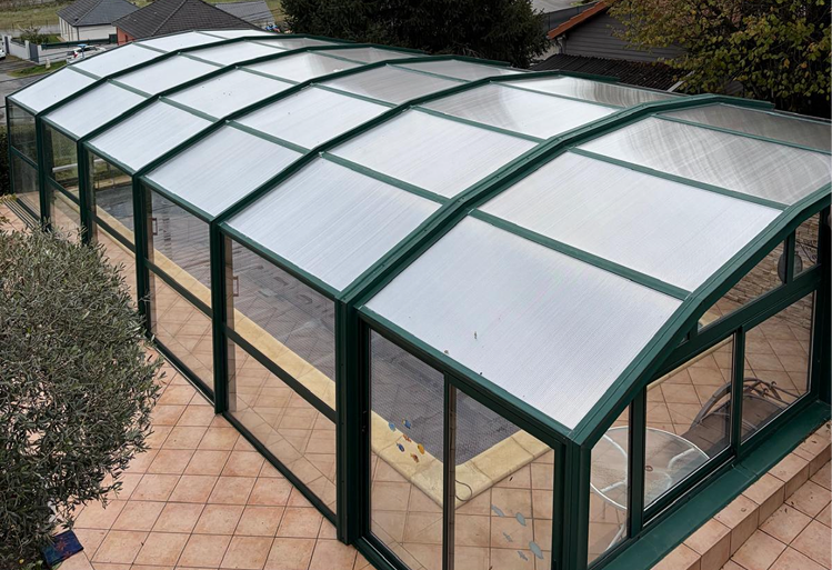 Grande véranda ou serre avec structure en métal vert et panneaux vitrés et toit ondulé translucide, installée sur une terrasse carrelée.