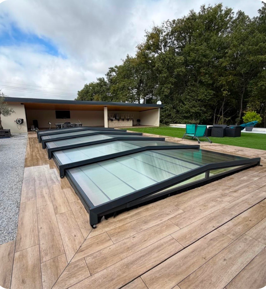 Toit de piscine rétractable en verre et cadre métallique noir installé sur une terrasse en bois près d'un espace extérieur avec pelouse, mobilier de jardin et arbres en arrière-plan.