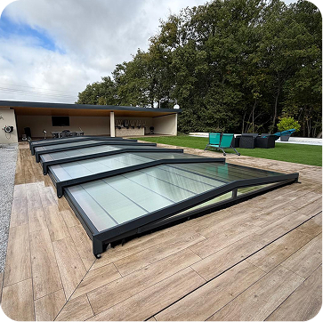 Piscine couverte par un toit rétractable en vitres et cadre noir sur une terrasse en bois, avec un salon de jardin et des chaises bleues sur une pelouse verte en arrière-plan sous un ciel partiellement nuageux.