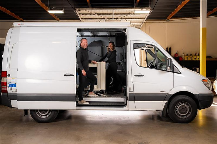 Two smiling people in black attire stand inside a white van for nuclear medical imaging. Industrial garage setting, tools in background.