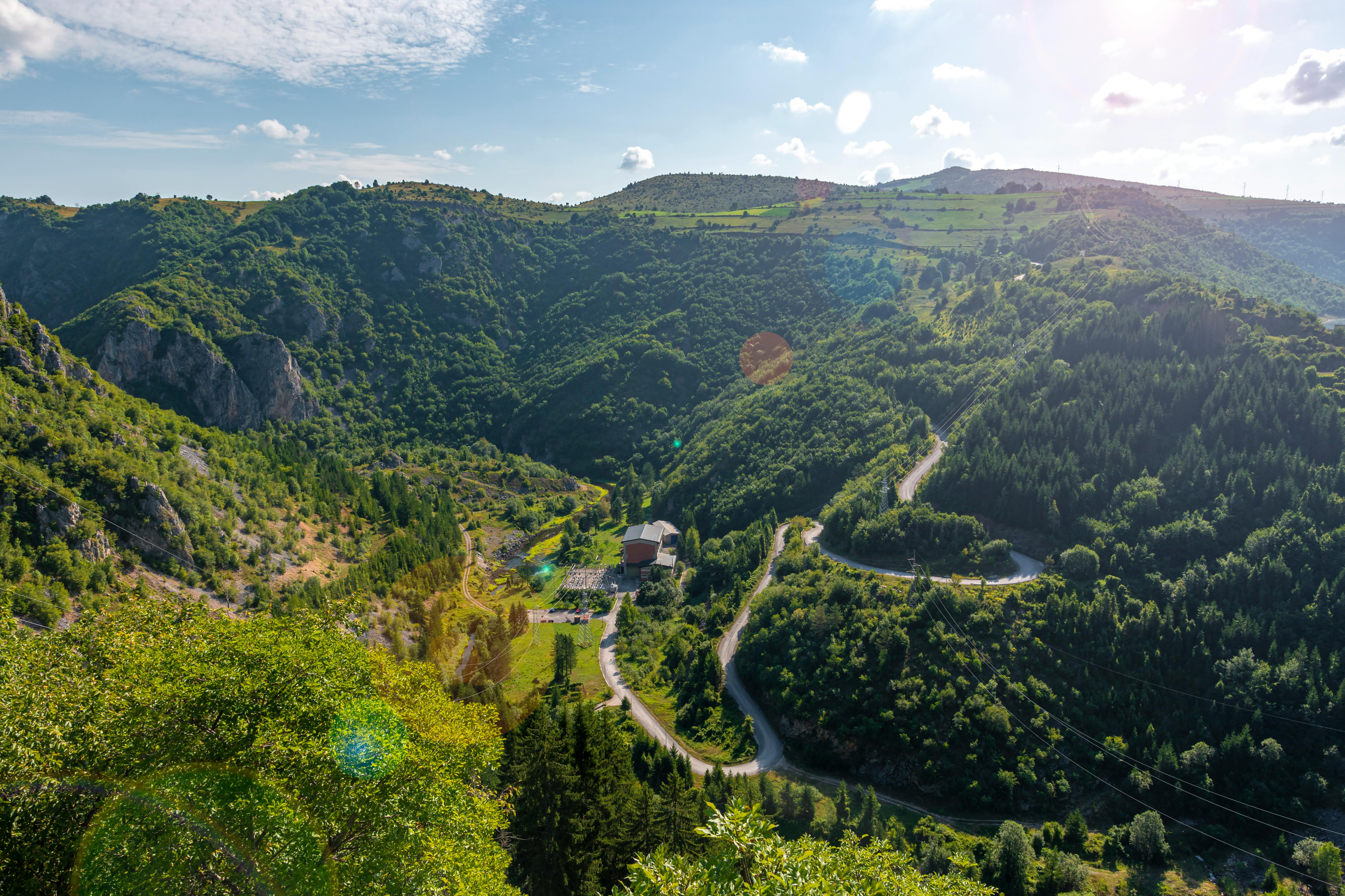 Winding road through lush green mountains with a building in a valley under a blue sky.