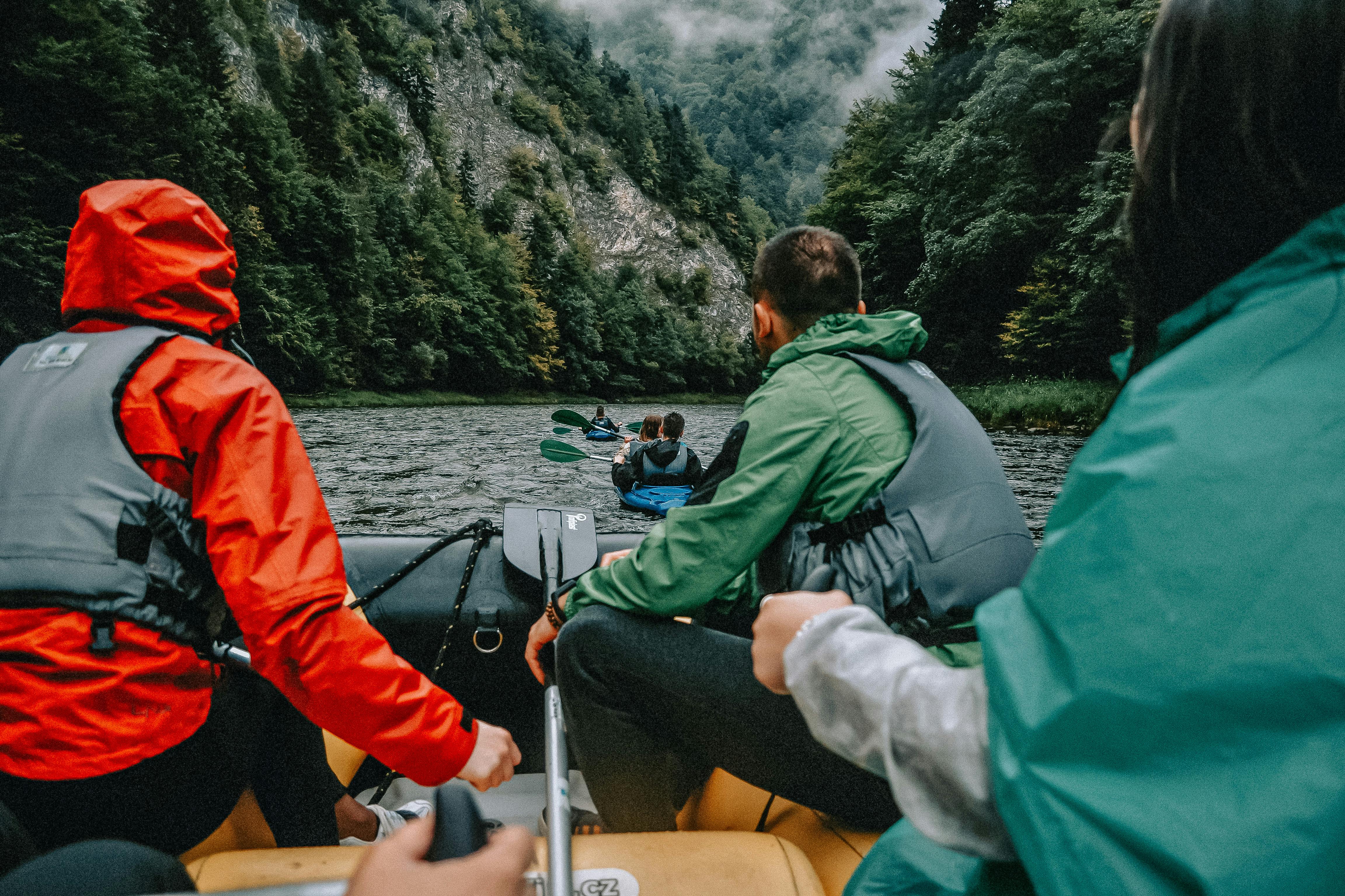 Three people in life jackets paddling a raft down a river surrounded by forested hills and mist.