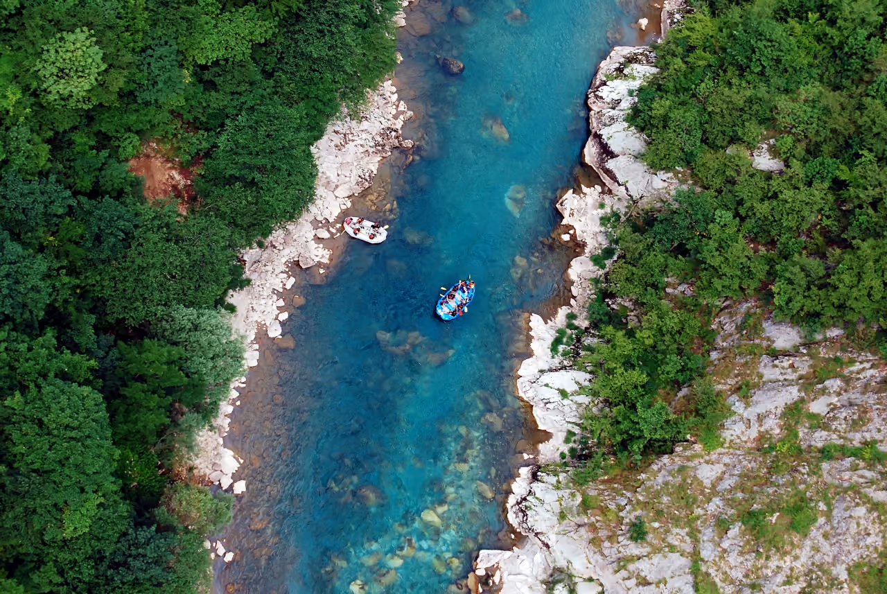 Aerial view of people rafting in a blue river surrounded by green forest and rocky banks.