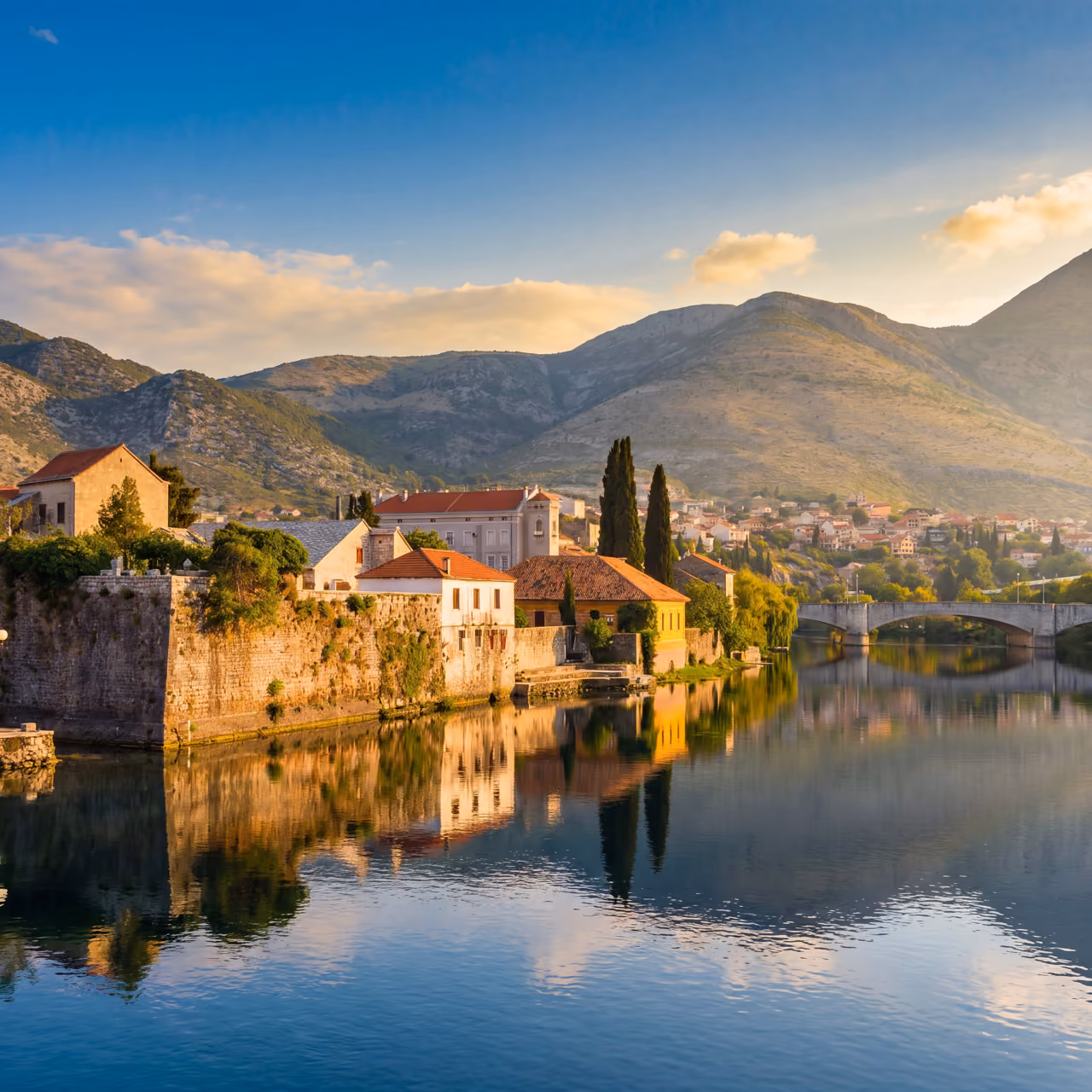 Old stone buildings with red roofs by calm river reflecting mountains and blue sky at sunset.
