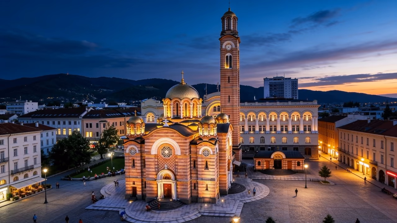 Lit Orthodox church with golden domes and tall clock tower in a city square at dusk.