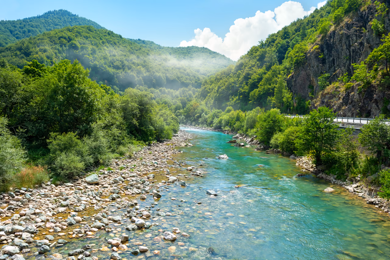 Clear blue river flows through forested mountains with rocky shores under a partly cloudy sky.