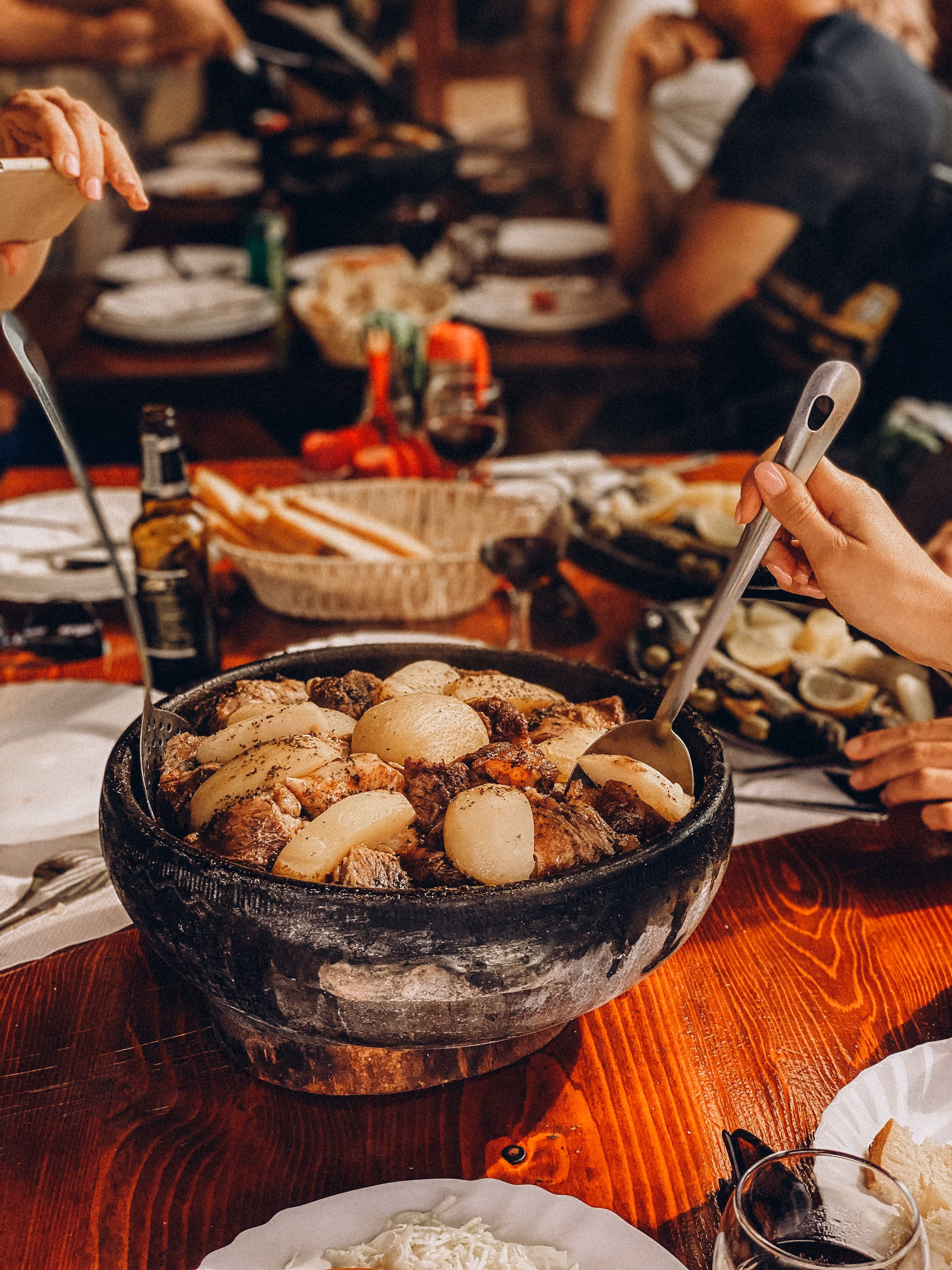 Hands serving a stew with meat and potatoes from a black pot on a wooden table at a meal.