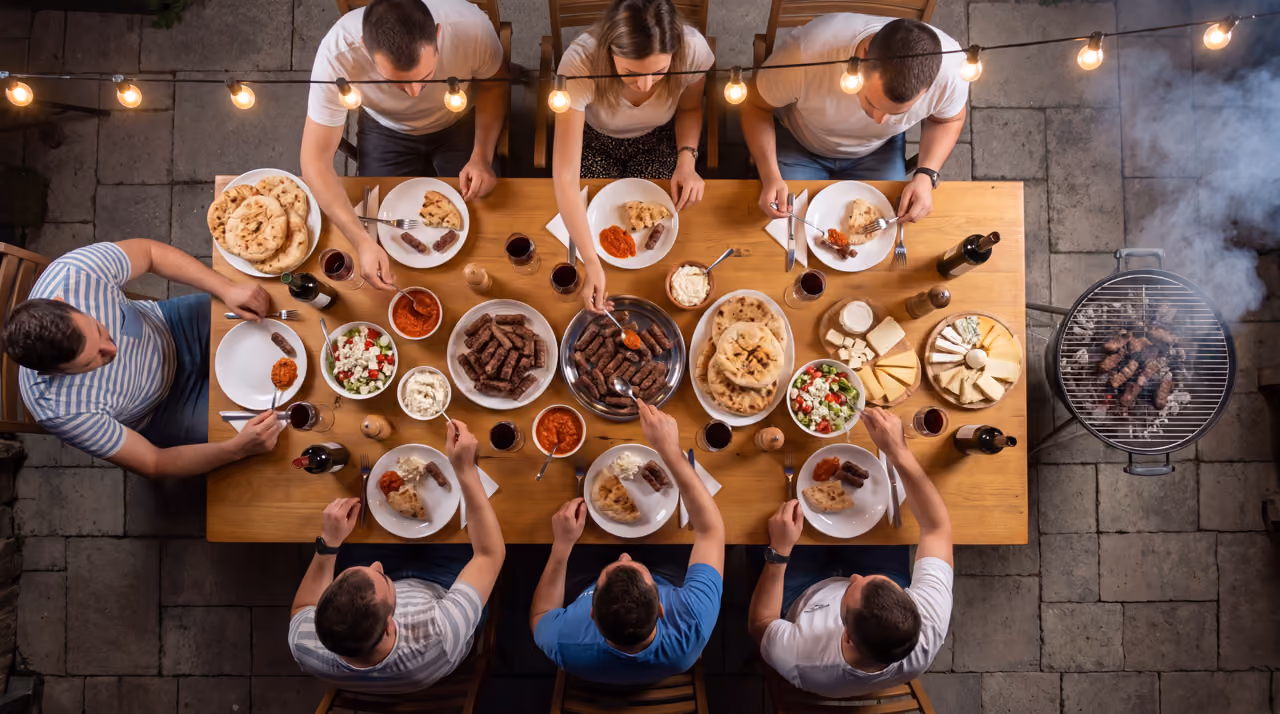 Overhead view of people eating grilled meat, flatbread, salad, and cheese at a wooden table outdoors.