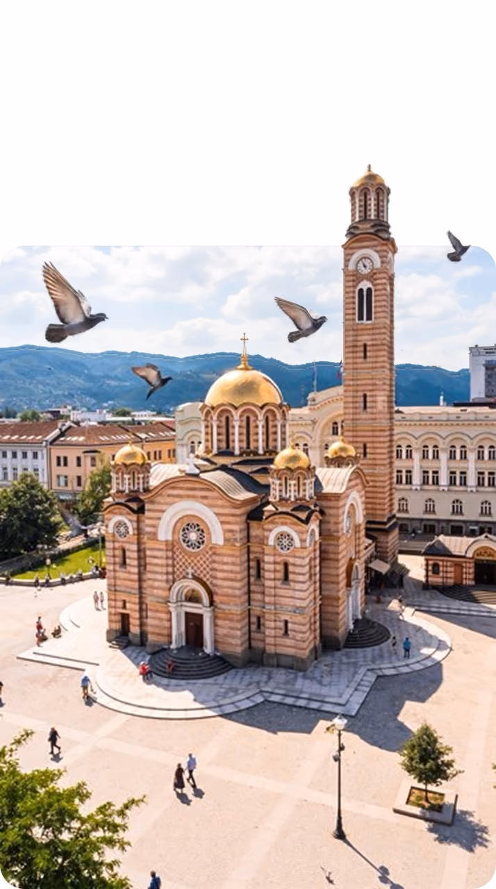 Orthodox church with golden domes and a tall bell tower in a plaza with birds flying overhead.