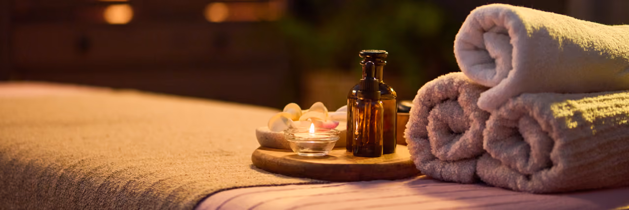 Rolled towels, a lit candle, and amber bottles on a wooden tray next to a massage table.