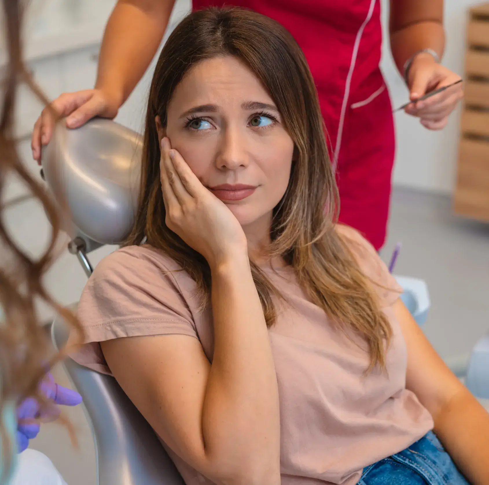 A woman sits in a dental chair, looking concerned and holding her cheek in discomfort.