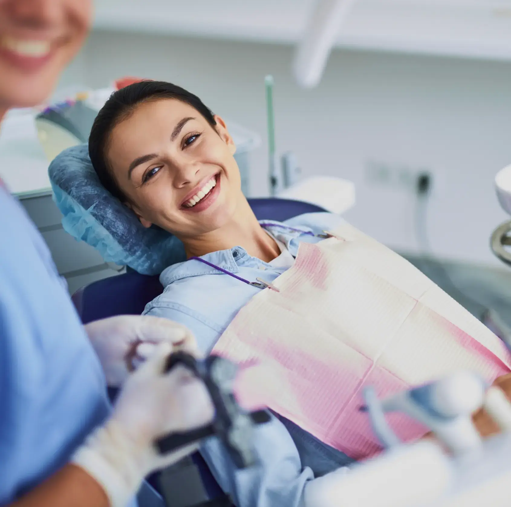 A woman sitting in a dental chair, smiling while a dentist prepares dental instruments.