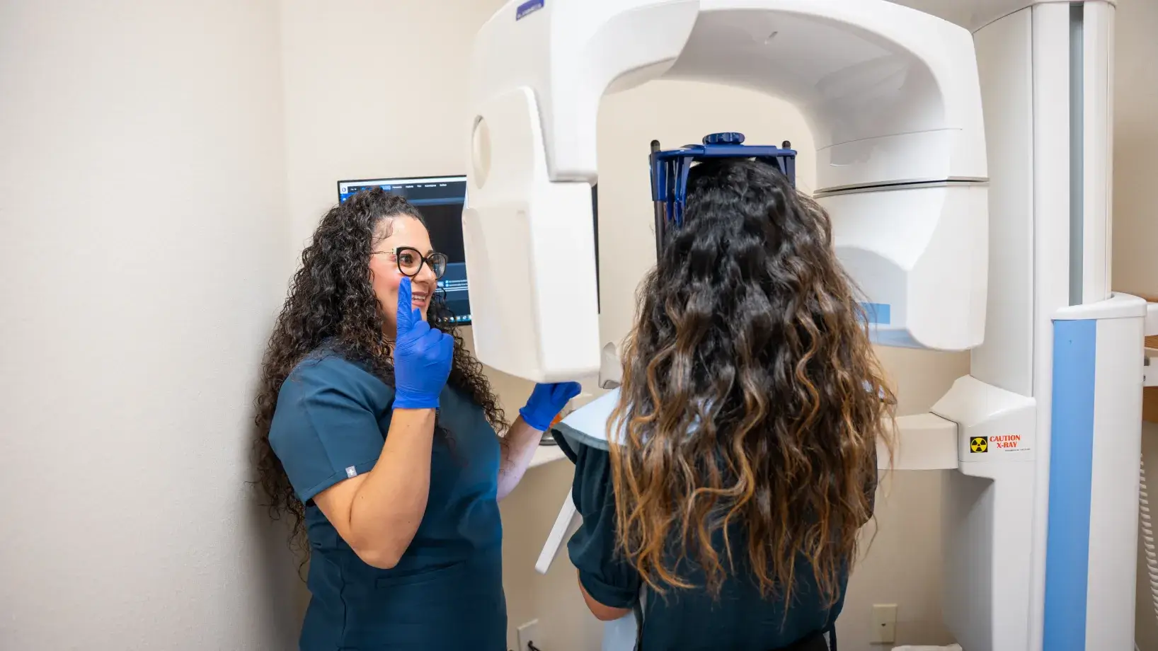 Two dental professionals prepare a patient for an X-ray scan using a large machine.