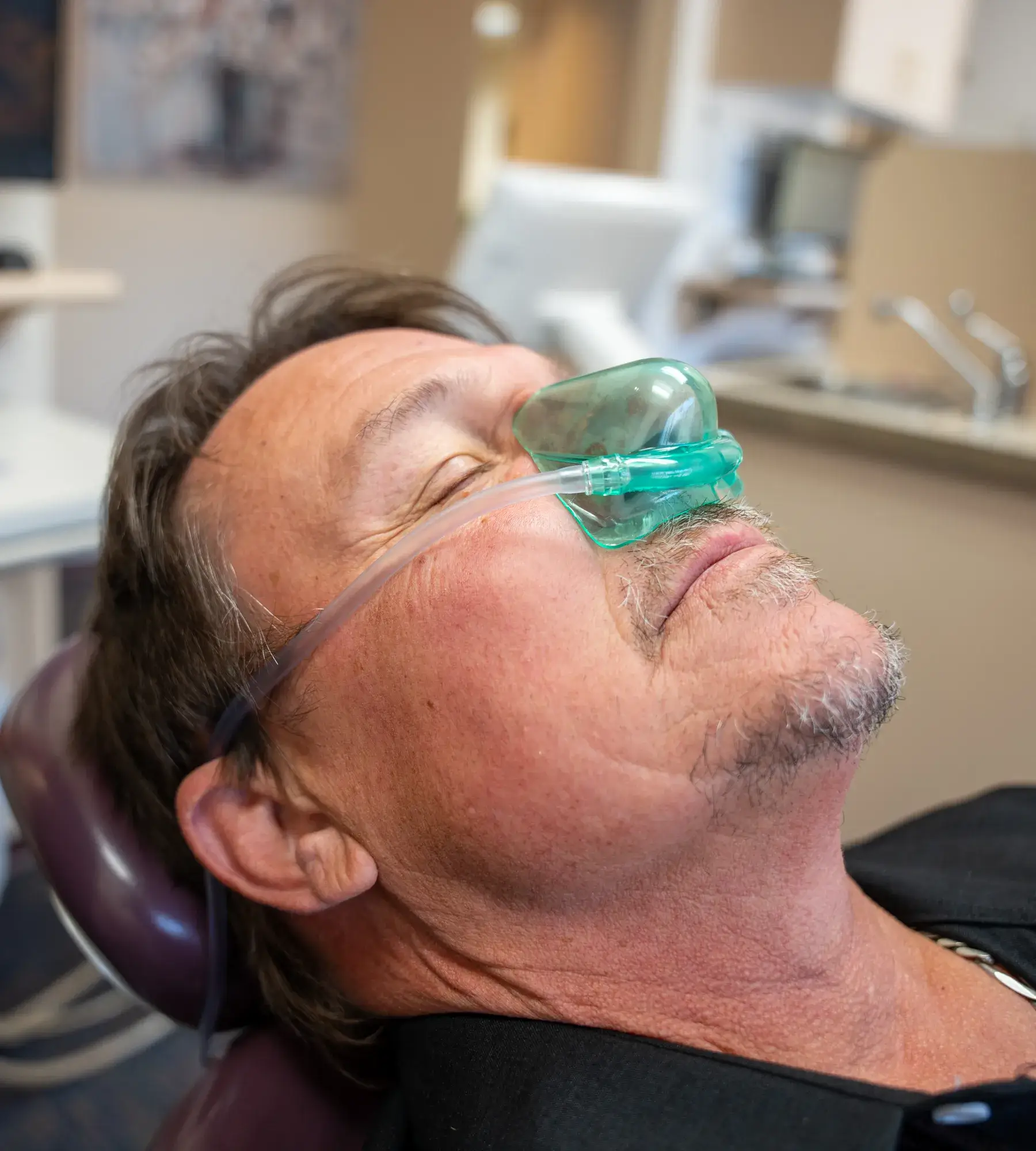 A man is reclining in a dental chair wearing a green nitrous oxide mask.
