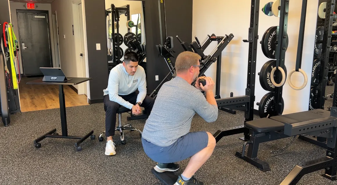 Man squatting with a weight ball while a trainer watches in a gym with fitness equipment.