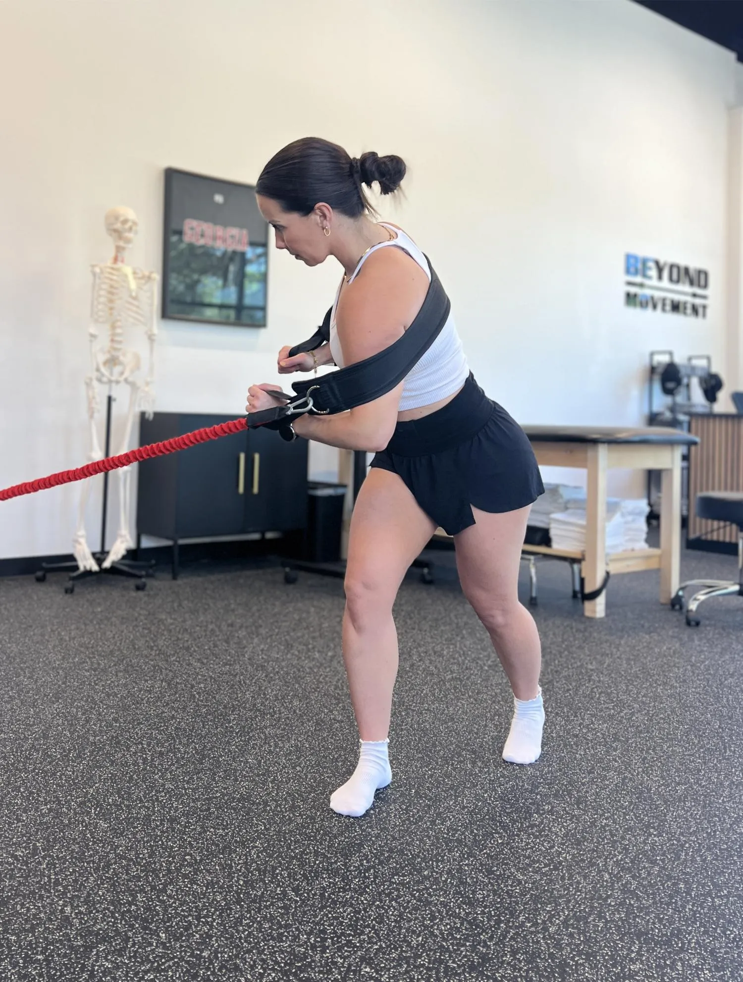 Woman in workout gear exercises using resistance bands and a shoulder harness in a gym with a skeleton model.