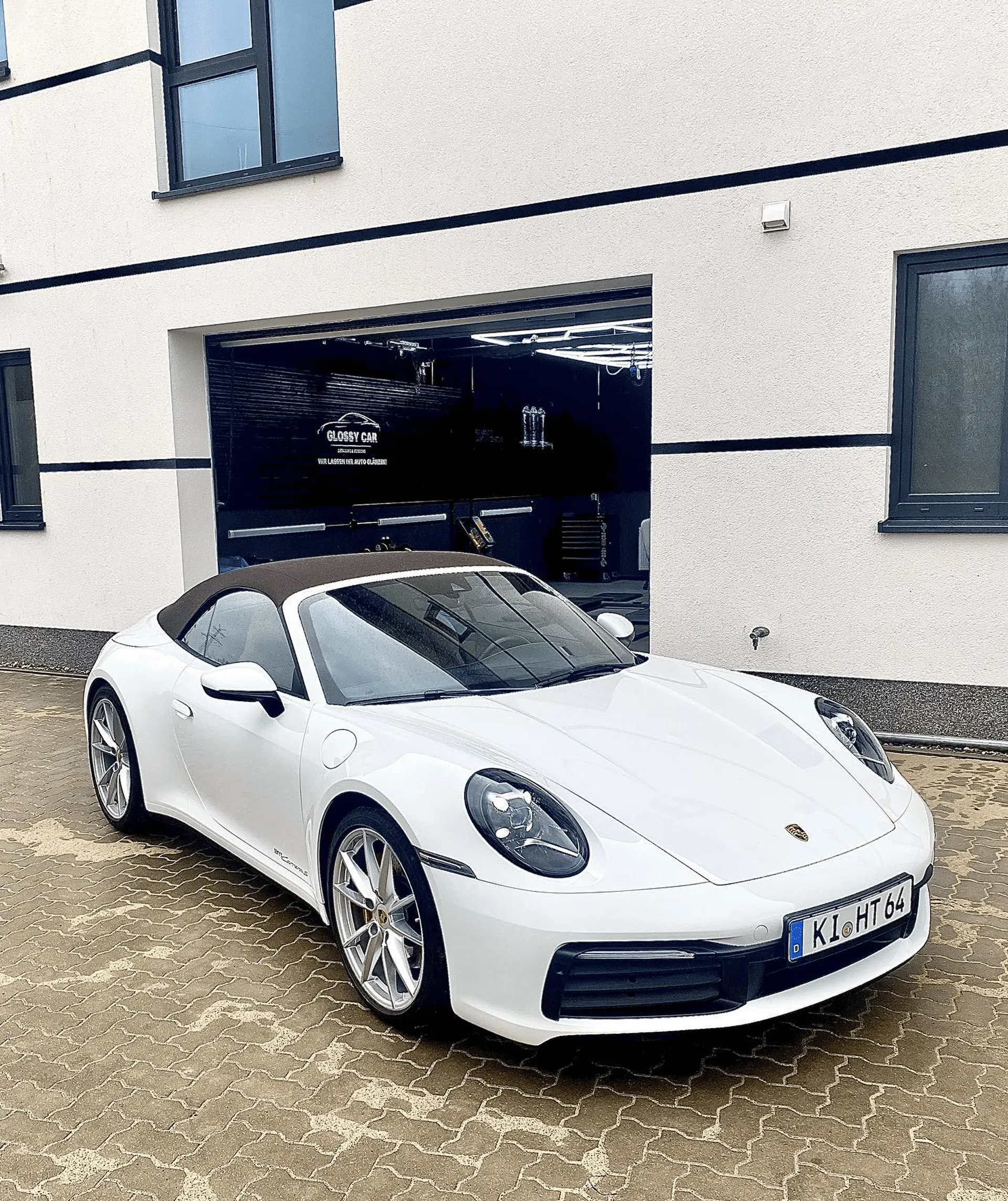 Weißes Porsche 911 Cabriolet vor der Garage von Glanzwerk Autoservice mit einem schwarzen Verdeck über dem Auto.