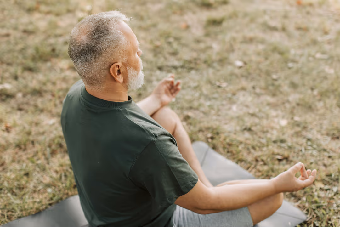 Older man with gray hair sitting cross-legged on a yoga mat outdoors meditating with hands in a mudra position.