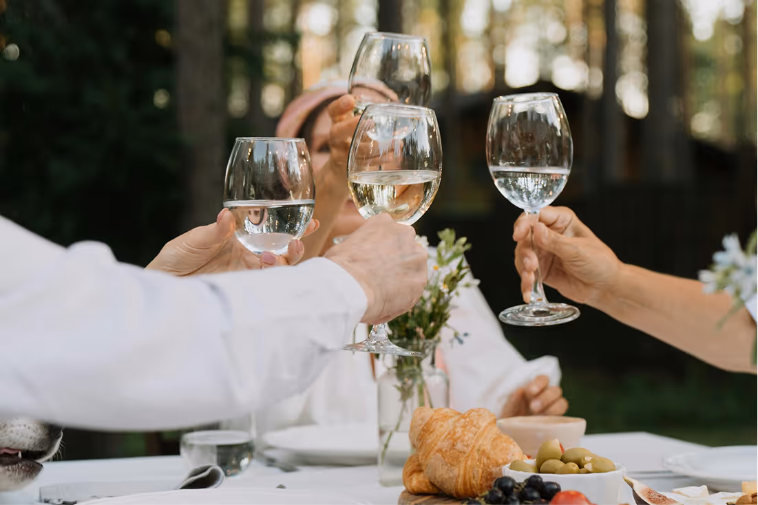 People clinking wine glasses over a table with croissants, grapes, and olives outdoors.