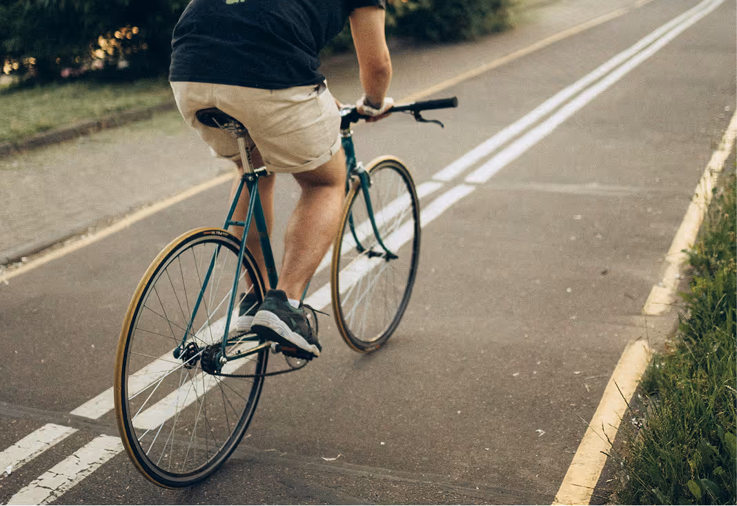 Person wearing shorts and a black shirt riding a teal bicycle on a paved path marked with double white lines.