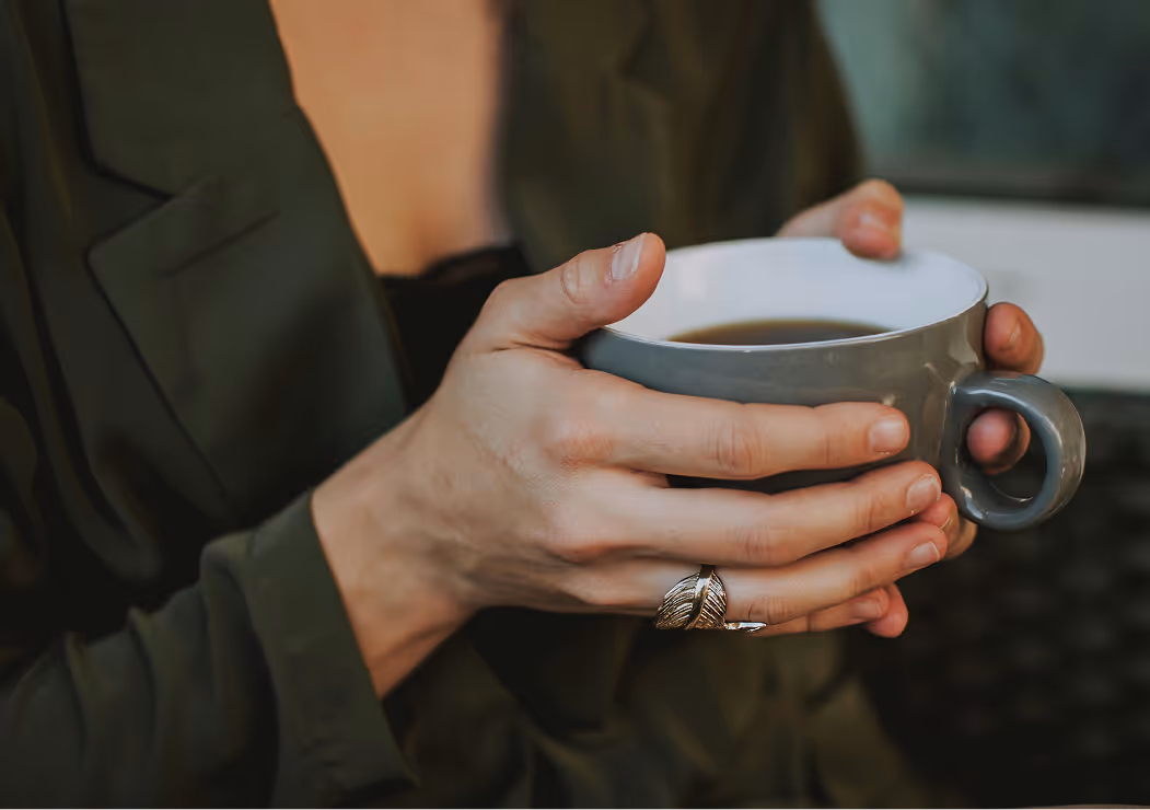 Person in dark clothing holding a gray cup filled with coffee with both hands, wearing a silver leaf-shaped ring.