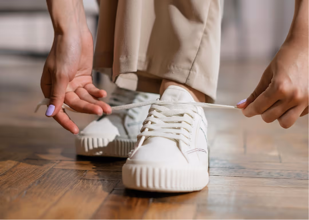 Person tying the laces of white sneakers while wearing beige pants on a wooden floor.