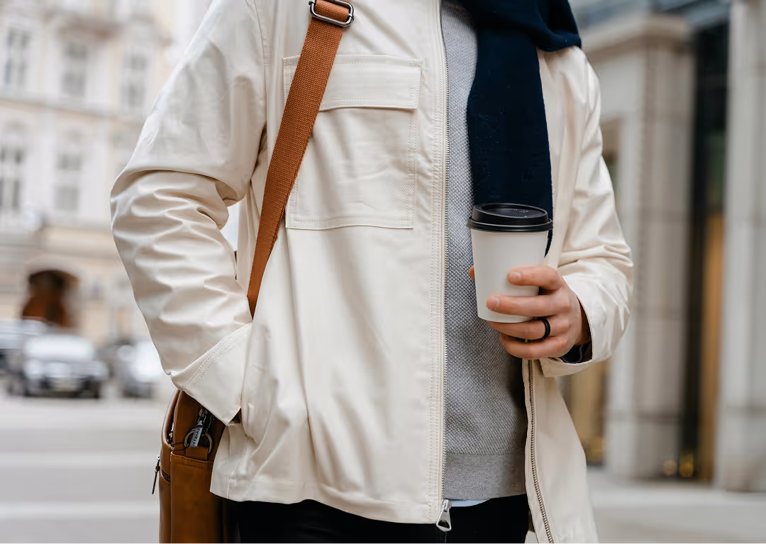 Person wearing a light jacket and scarf, holding a disposable coffee cup, with a brown shoulder bag.