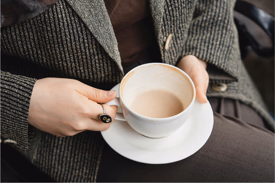 Person wearing a textured gray coat holding a white cup and saucer with light brown coffee.