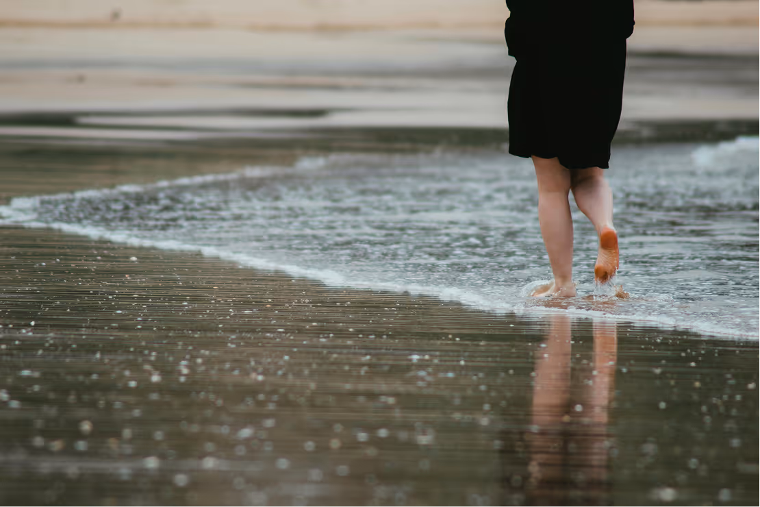 Person walking barefoot along the shoreline with water washing over sand.