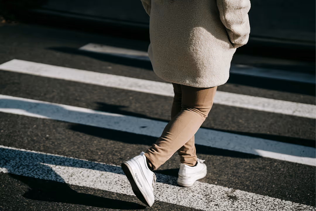 Person wearing a beige coat, brown pants, and white sneakers walking across a crosswalk.